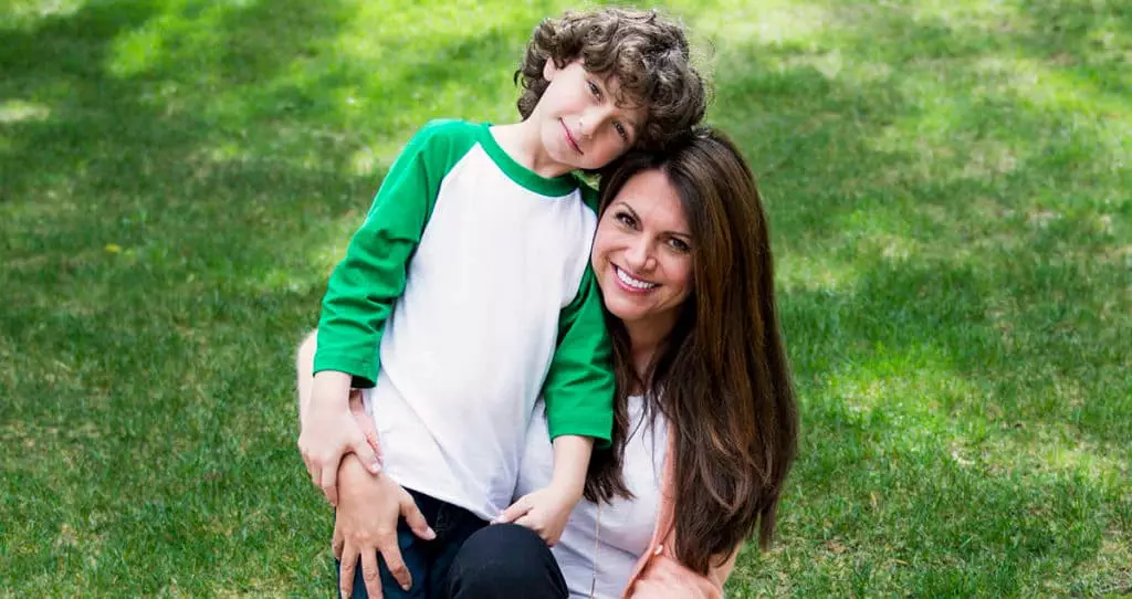 August "Auggie" Maturo and his mom. He is leaning into her and she's holding him. They are sitting outside on wooden steps with grass in the background.