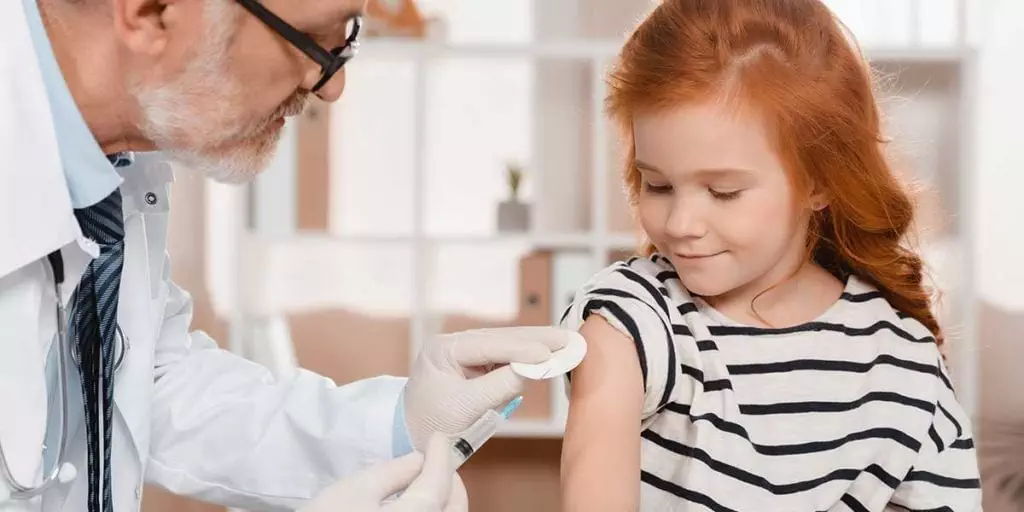 Photo of male doctor vaccinating a child in her arm. She looks on with a small smile and is not scared.