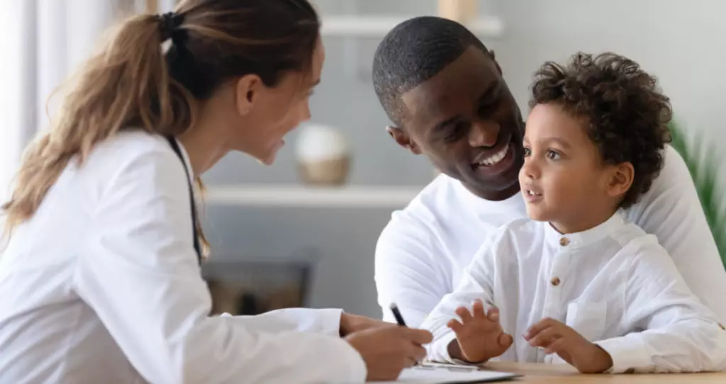 Cheerful young female pediatrician and african american smiling father listening to mixed race little patient, telling doctor about well-being. Happy multiracial family visiting clinic for check up.
