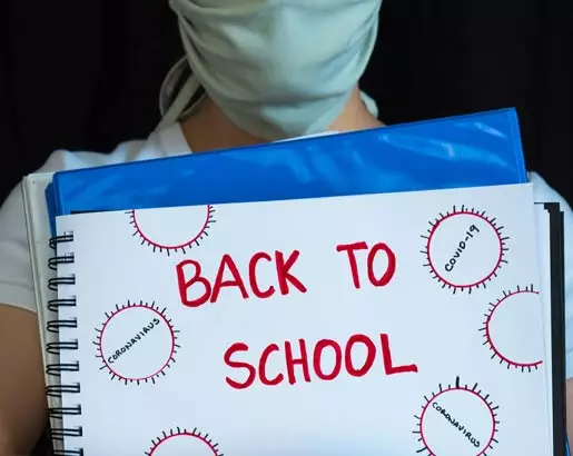 Image of teach with medical mask on her face holding a notebook that says "Back to school." The notebook has coronavirus images that are hand drawn on it.