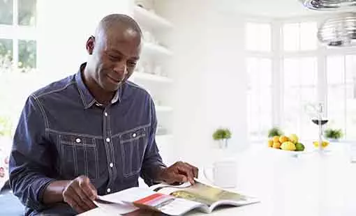 Image of a Black man reading at his kitchen table. He looks interested in the information he is reading