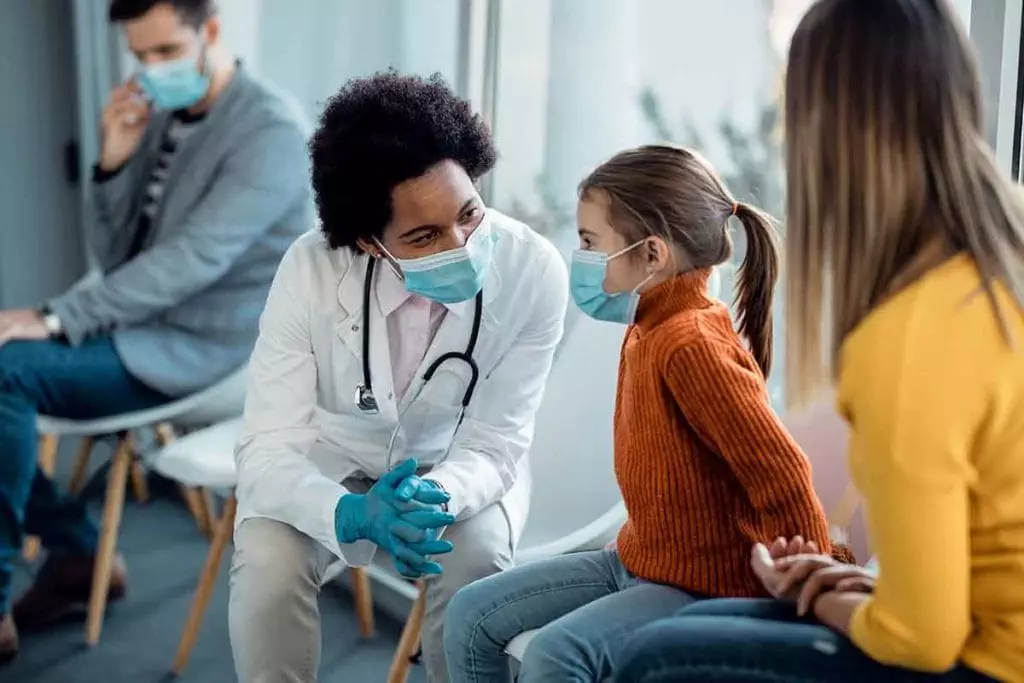 Photo of a doctor wearing a mask talking to a child patient wearing a mask