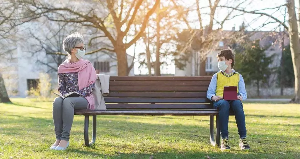 Photo of grandmother and grandson visiting with each other. They are sitting far apart on a park bench and they each are wearing masks. The grandmother has asthma and reduces her stress by safety visiting with her family.