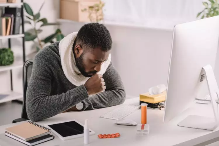 Black man sitting in his home office in front of a computer. He is coughing into his hand and there are signs of medication on the desk. The man has whooping cough as an adult.