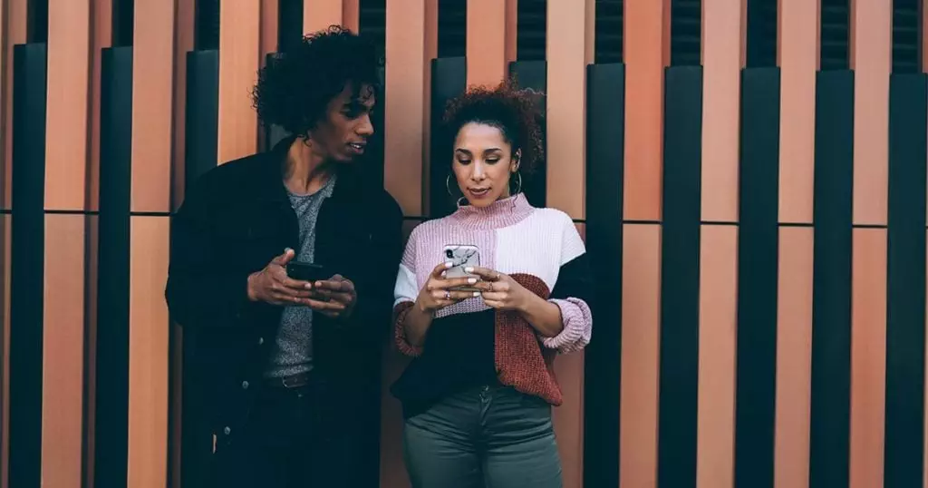 Photo of a black man and woman , against a wall, looking at their phones