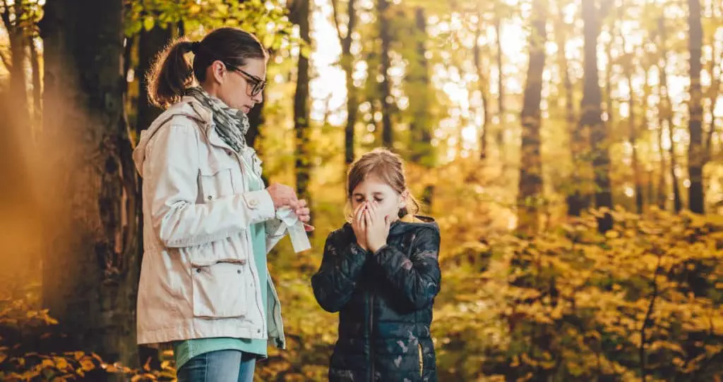 Child is sneezing from the hay fever pollen in the park. Her mother is standing next to her holding a tissue, waiting to hand it to her. It appears to be the autumn season.