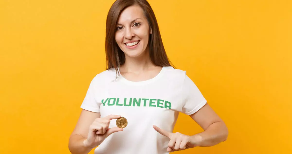 Woman in white Tshirt with the word "volunteer" on it is holding up a bitcoin coin and pointing to it with her other hand. The concept is the organization accepts cryptocurrency donations including Bitcoin.