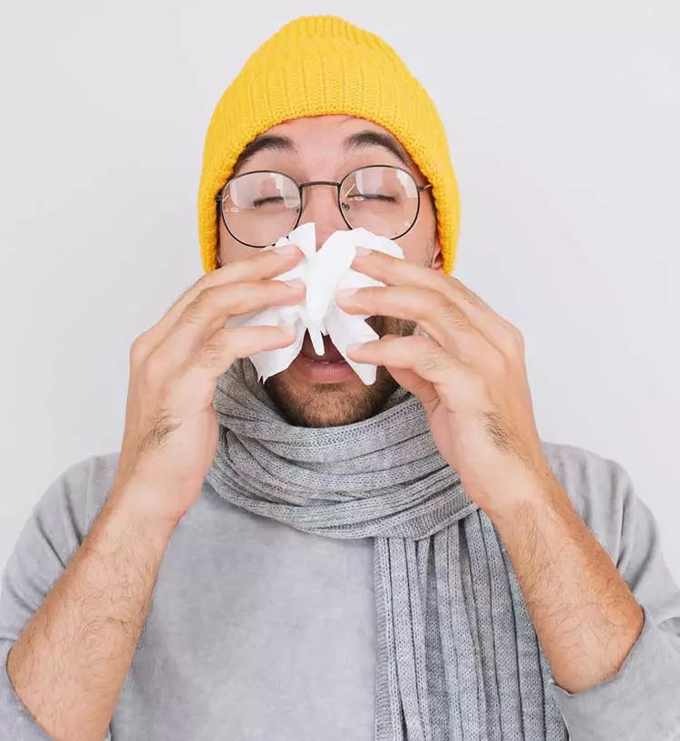 Photo of a young man in a yellow hat and grey shirt. He is holding a tissue to his nose as he's about to sneeze.