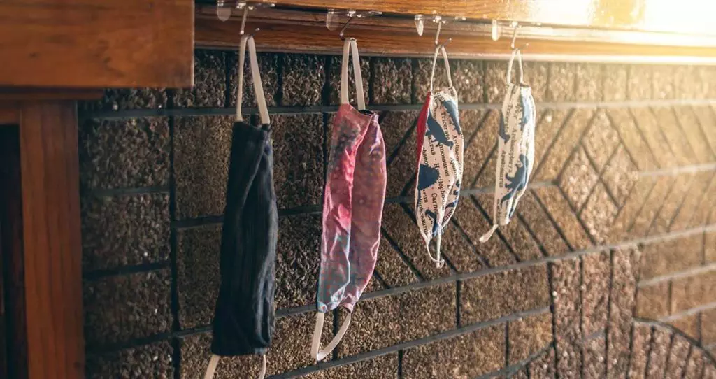 Photo of various size homemade face masks hanging on a coat rack that is mounted to a wall.