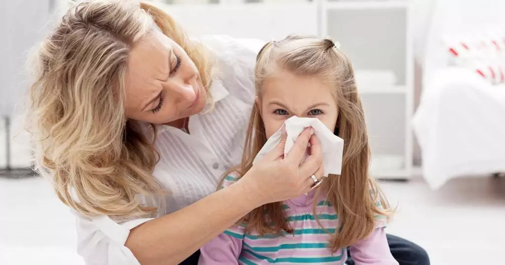 Photo of white mother with her child, in the child's bedroom. She is holding a tissue to the child's nose because of allergy symptoms.