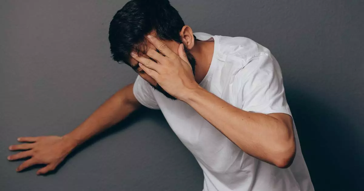 Man in white Tshirt holding his head and  leaning against a wall showing signs on an anaphylaxis episode