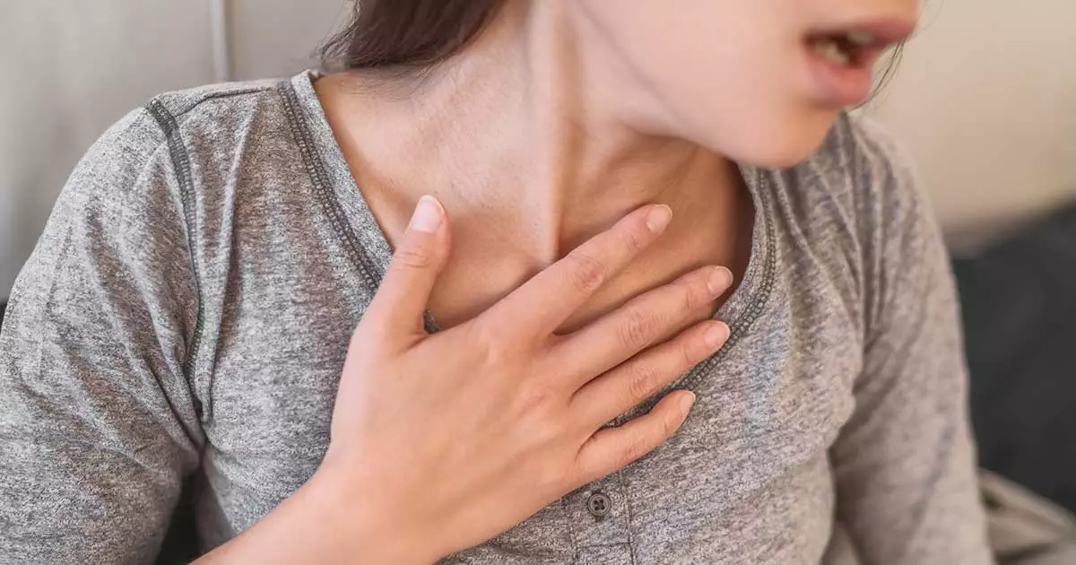 Young adult woman in grey shirt holding her hand to her throat and struggling to breathe because she's having an anaphylaxis episode