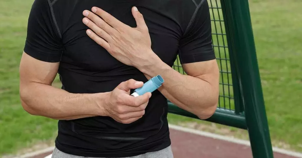View of the torso of a fit man in a sport park. He is holding an inhaler and grasping his chest.
