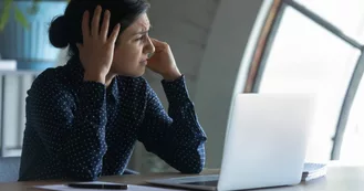 Photo of a south eastern Asian woman sitting at a desk holding her head with a migraine. She is wearing a dark long sleeved shirt with laptop on the desk in front of her.