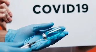 Photo of latex gloved hands with needle inserted into medication vial and COVID-19 in text in the background