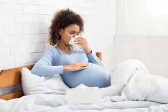 Pregnant Black woman laying in bed holding a tissue to her nose. She appears to have allergies.