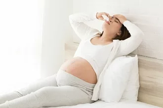 Pregnant woman laying on the bed holding her head up because of allergies.