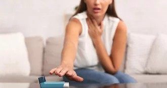 Photo of young adult woman sitting on a couch. She is holding her neck are with one hand and reaching to grab her asthma inhaler with the other hand.