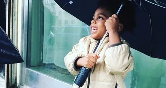 Three year-old Elijah Alavi Silvera holding an umbrella while outside in front of a window. He has a smile on his face and he is looking up.