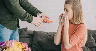 White woman with blonde hair sitting on the couch and holding a tissue to her nose because she is sneezing. There is man standing next to her with a glass of water and allergy meds he is handing to her.