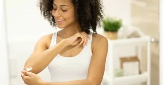 Young Black woman with a smile, wearing a tank top, isputting eczema cream on her elbow