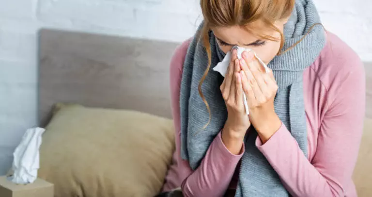 Photo of woman with covid and asthma. She is blowing her nose into a tissue and has a grey scarf wrapped around her neck. There is a tissue box next to her.