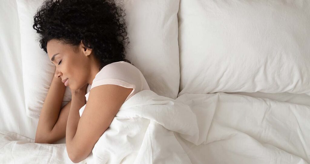 非裔美国妇女睡在舒适的床上，躺在柔软的枕头矫形床垫上，安静的年轻黑人女士躺在卧室的白色床单上，盖着毯子，俯视图